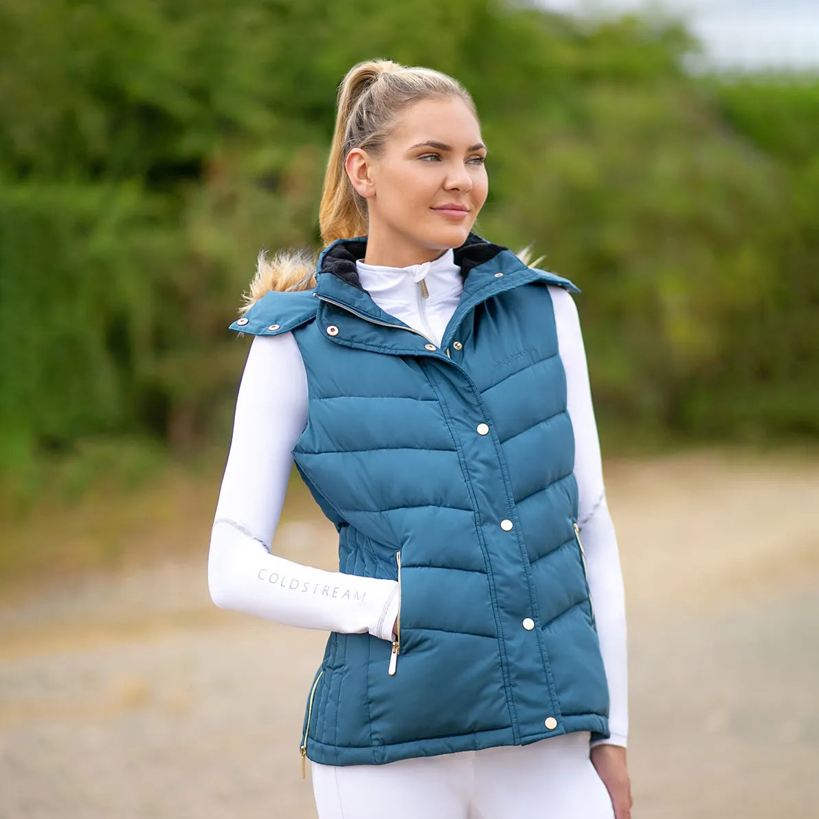 Woman wearing a teal puffer vest outdoors with a blurred green background