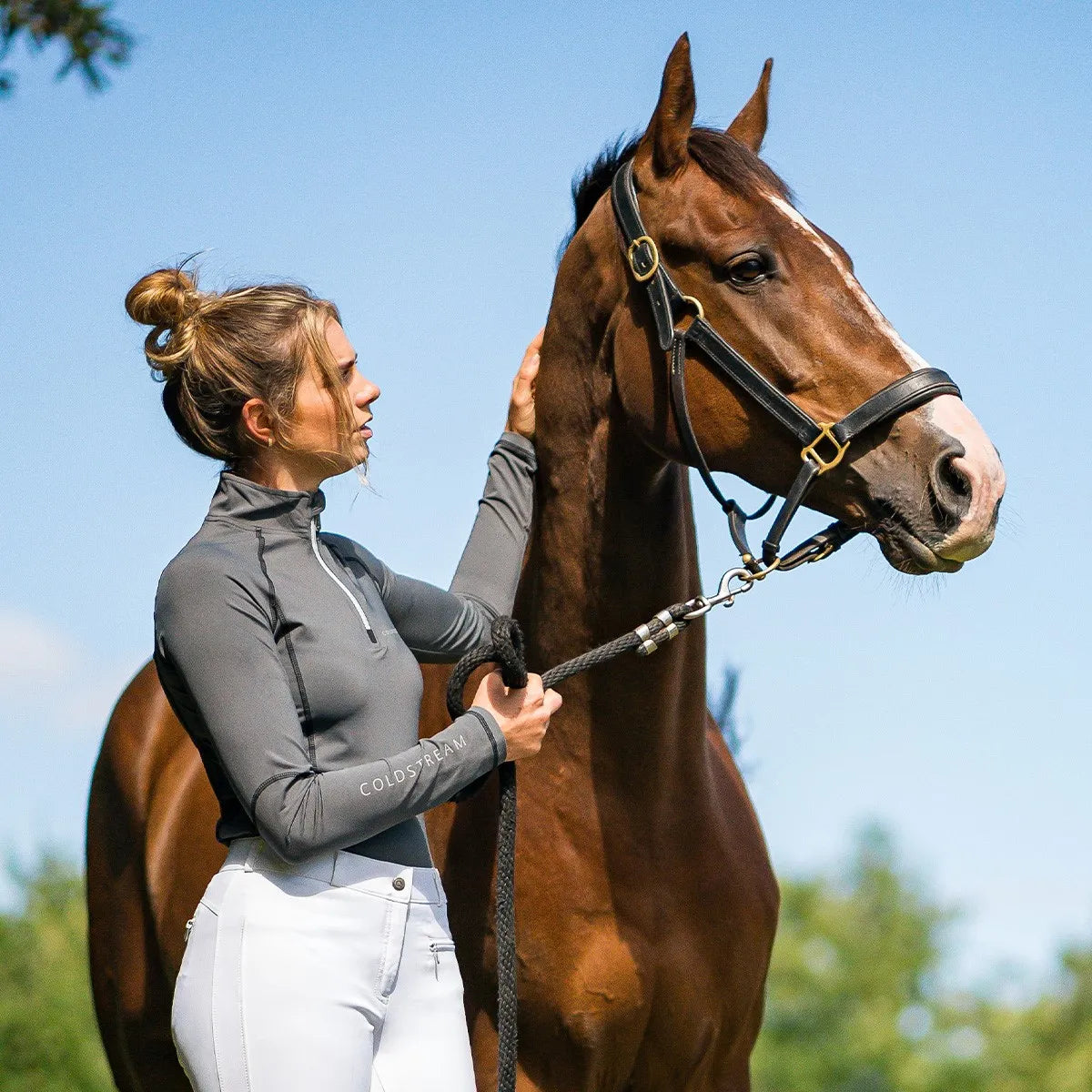 Woman leading a brown horse outdoors with a clear blue sky.