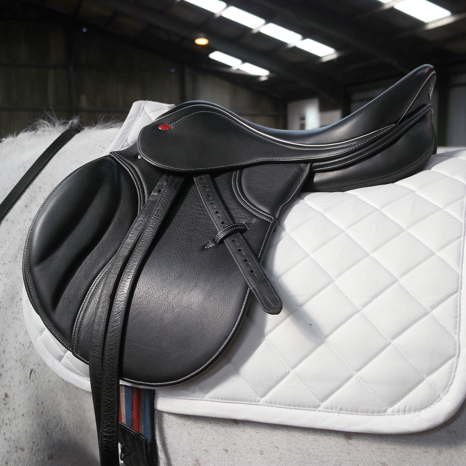 Black leather saddle on a white quilted saddle pad in an indoor equestrian setting.