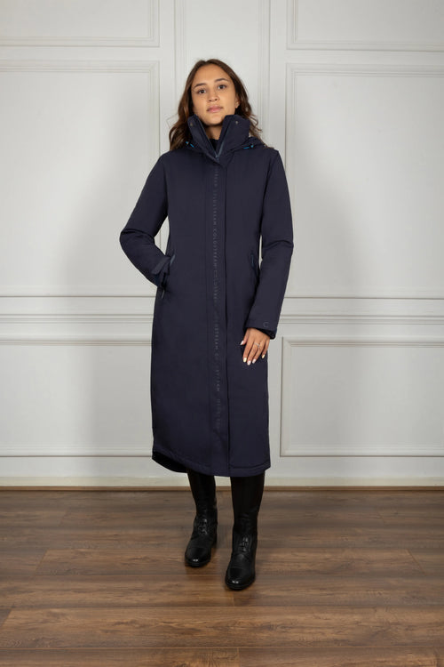 Woman wearing a long navy coat against a white paneled wall.